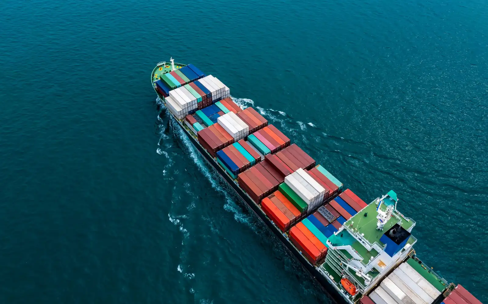 An aerial shot of a barge transporting multi-coloured shipping containers on a deep cerulean sea.